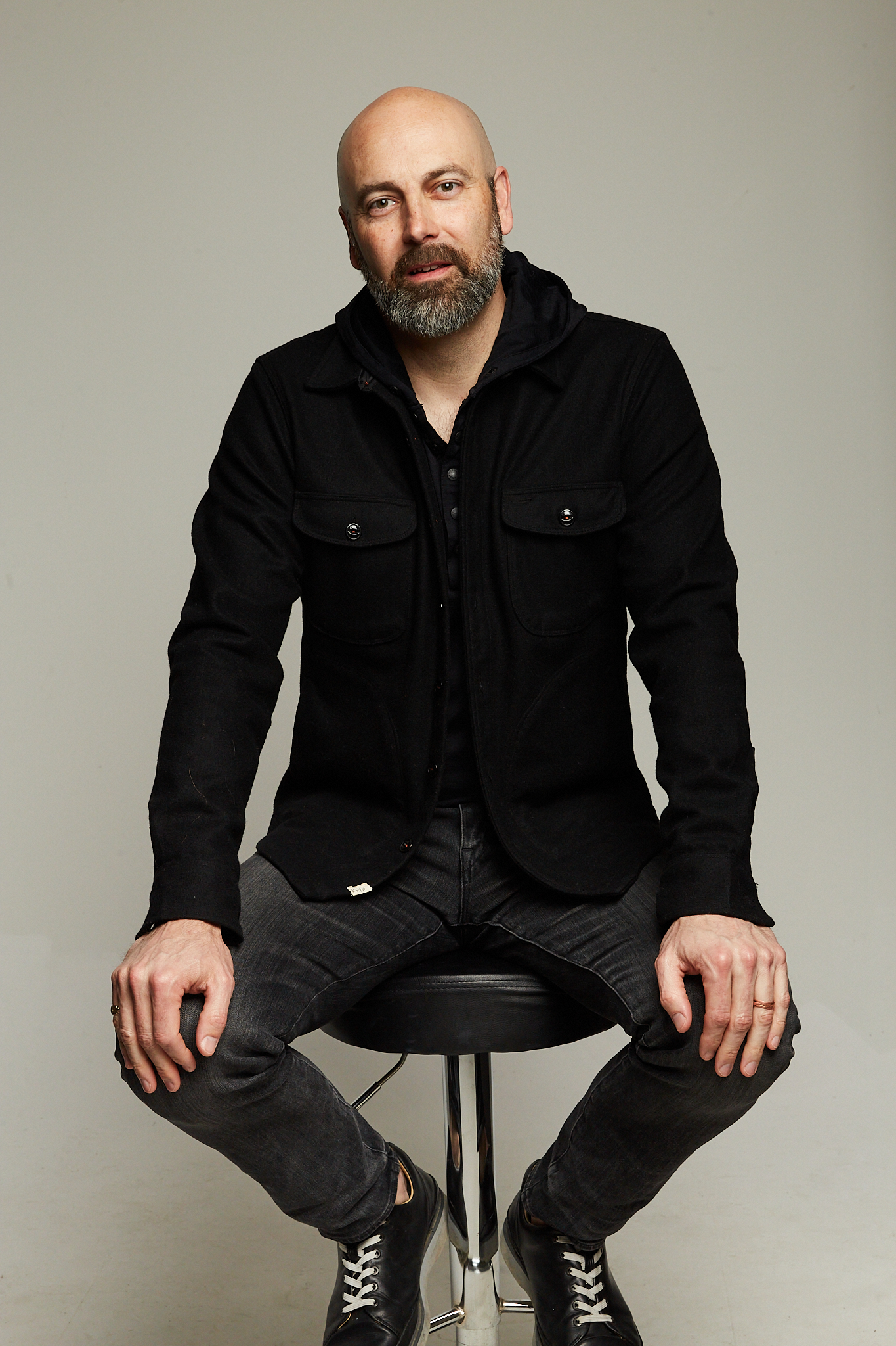 Ben Callahan, seated on a stool, dressed nicely in all black. He has a slightly graying beard and a shaved head, a tall thin build and a curious look in his eyes.